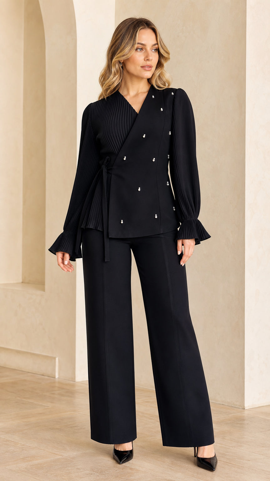 Full front view of model wearing black pleated wrap blouse with tie waist and pearl accents