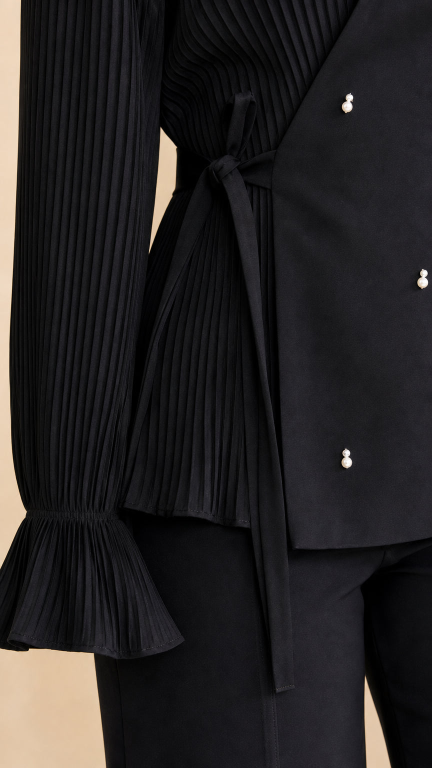 Close-up detail of black blouse showing accordion pleats, pearl beads, and tie waist