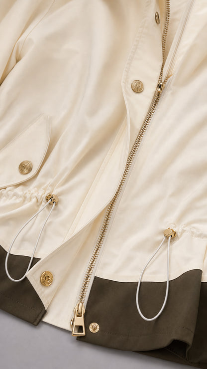 Close-up detail of cream jacket showing gold zipper, drawstring waist toggles, snap button, and olive contrast hem