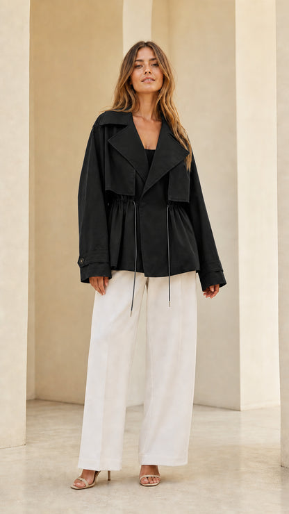 Front view of model wearing black short trench jacket with white trousers