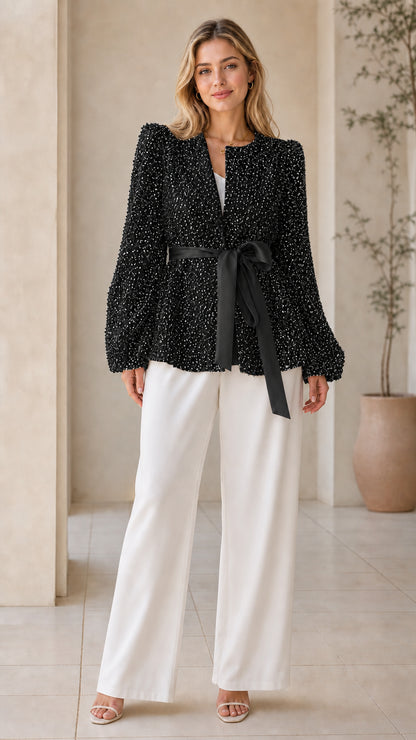 Front view of woman wearing black sequin tie-waist party jacket with white trousers